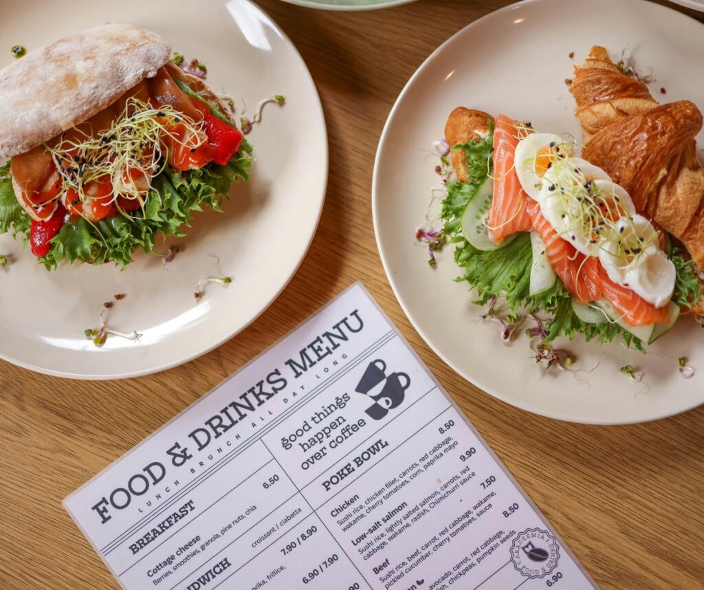 Two plates with open sandwiches (one with salmon and egg beside croissants) on a wooden table next to a ‘Food & Drinks Menu.