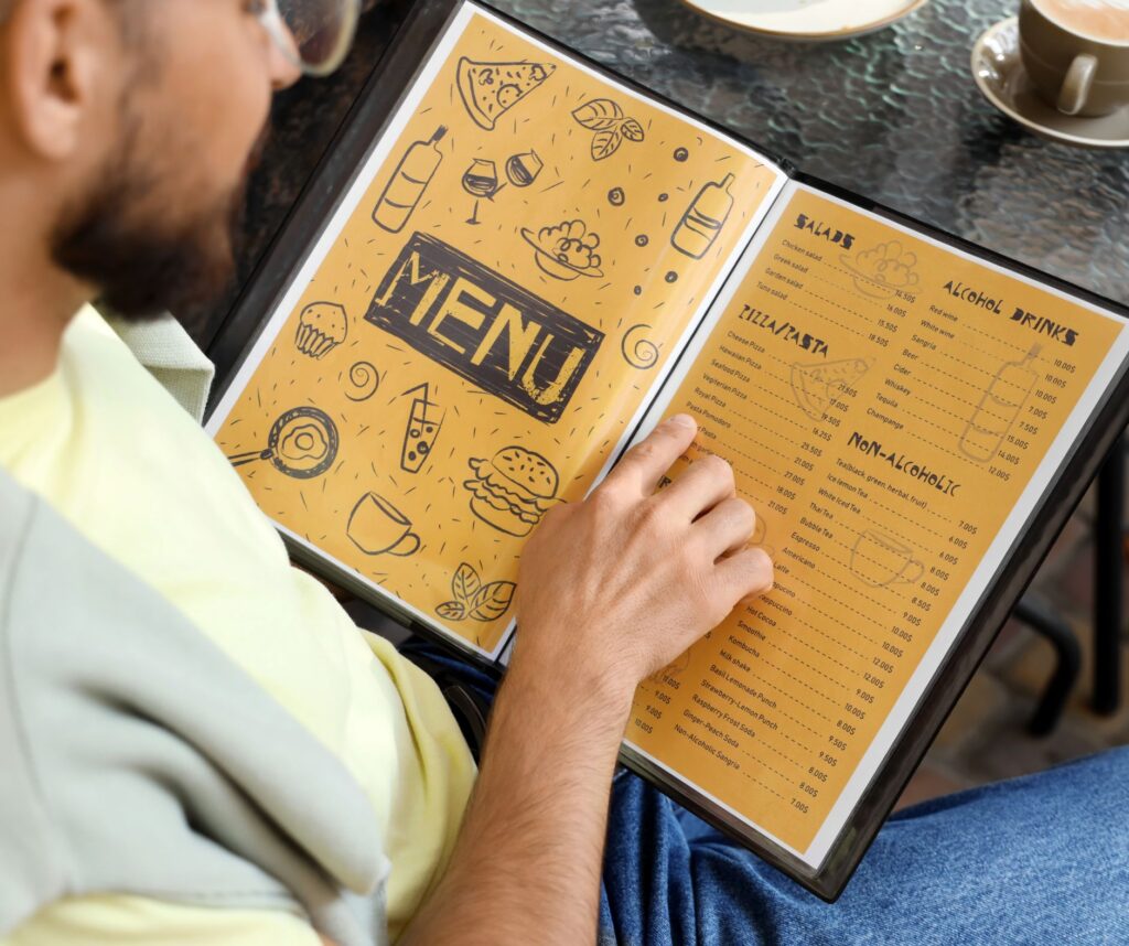 Person seated at a table holding open a restaurant menu with a yellow illustrated cover, with cups and drinks on the table nearby