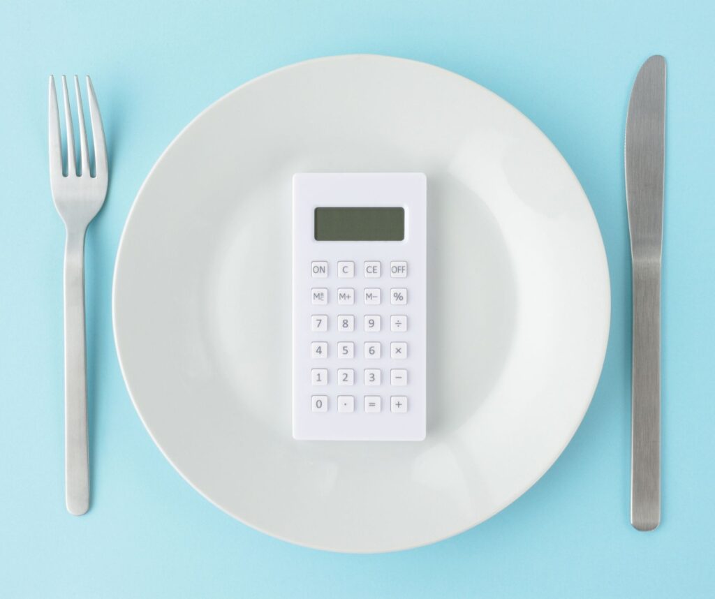 Calculator on a dinner plate between a fork and knife, symbolizing budgeting, portion control, and food cost savings.